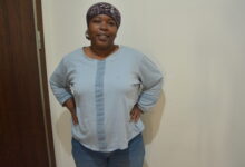Marie Gunduz in a blue shirt, blue jeans, at 101 kg with a head scarf on looking at the camera. Standing next to a white wall and brown door.
