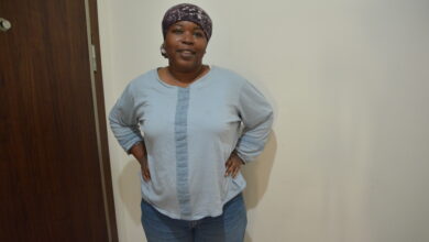 Marie Gunduz in a blue shirt, blue jeans, at 101 kg with a head scarf on looking at the camera. Standing next to a white wall and brown door.