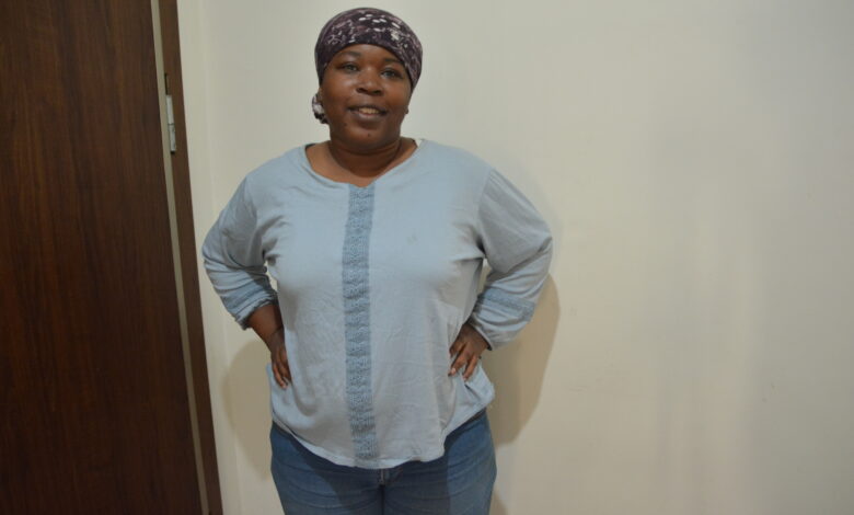 Marie Gunduz in a blue shirt, blue jeans, at 101 kg with a head scarf on looking at the camera. Standing next to a white wall and brown door.