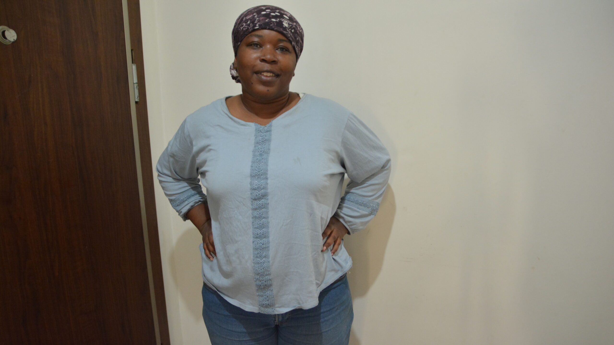 Marie Gunduz in a blue shirt, blue jeans, at 101 kg with a head scarf on looking at the camera. Standing next to a white wall and brown door.